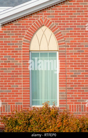 Arched window of a church with red brick wall. Close up view of the arched window of a church with red brick wall on a sunny day. Lush green plants ca Stock Photo