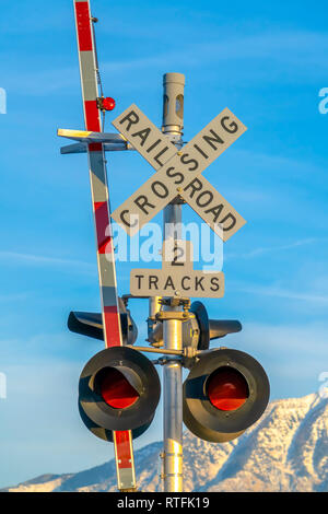 Railroad crossing sign with barrier and red lights. A two tracks railroad crossing sign against snow caped mountain and blue sky. A railroad barrier a Stock Photo