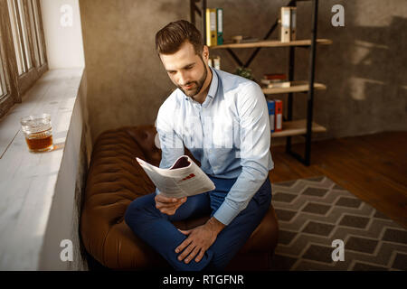 Young handsome businessman reading journal in his own office. He sit at window and read journal. Glass of whiskey stand on windowsill. Sunlight on wal Stock Photo