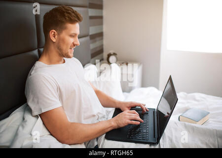 Cheerful nice young man work in bed early moring. Guy hold laptop and type on keyboard. Smile and positive. Daylight Stock Photo