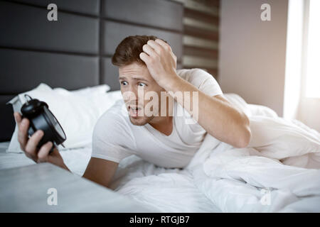 Emotional young man on bed in early morning. He overslept. Guy hold clock and look at it scared Stock Photo