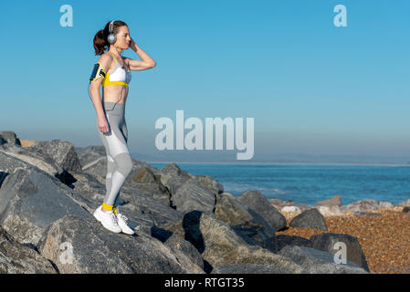 woman wearing sportswear standing on rocks looking out to sea listening to music on her smartphone, relaxing after exercise Stock Photo