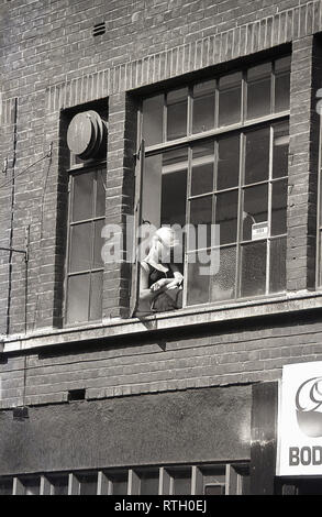 1970s, Summertime in the city and a young female office worker taking a break, sitting reading at a open window in an old building in East London, England. Stock Photo