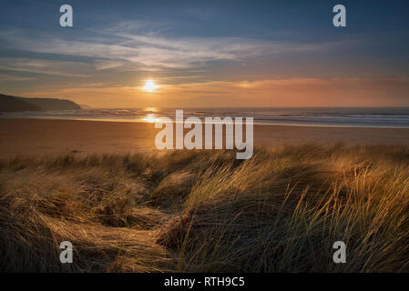 Golden sun spreading across the wide sand expanse of Perranporth beach and the marram grass on the sand dunes at sunset on the west cornwall coast Stock Photo