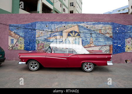 Classic 1960's American car parked against a colourful wall in Cuba with 1960's buildings in distance Stock Photo