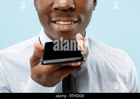 Indoor portrait of attractive young black african man isolated on blue background, holding blank smartphone, using voice control, feeling happy and surprised. Human emotions, facial expression concept. Stock Photo