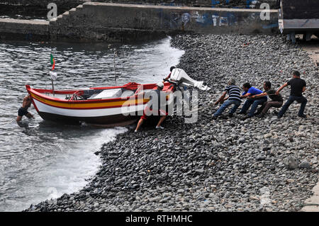 Around Madeira - Fishermen at the end of a long day, pulling their boat up onto a pebbled beach at Camara de Lobos. Stock Photo