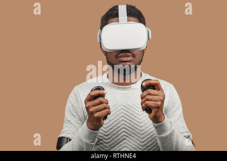 Dark-skinned millennial tourist in casual clothing wearing glasses with head-mounted display for smart phone, operating with controllers in hands, doi Stock Photo