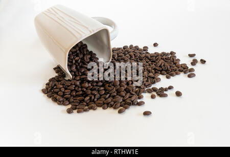 White, porcelain coffee cup tipped over with whole roasted coffee beans spilling out on a white background Stock Photo