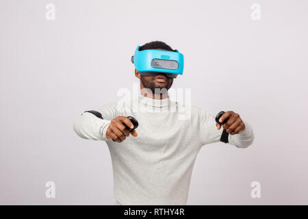 Dark skinned ethnic man using virtual reality glasses watching a simulation image 3D, holding gamepads in raised hands as if he plays Car Race Stock Photo