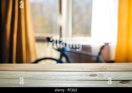 Product Placement Background. Wooden Table Against Blurry Interior of Cyclist's Living Room with Large Window and Blue Sport Bicycle Stock Photo