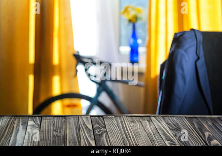 Product Placement Background. Wooden Table Against Blurry Interior of Cyclist's Living Room with Large Window and Blue Sport Bicycle. Blue Jacket on t Stock Photo