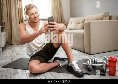 Young well-built man go in for sports in apartment. Happy positive guy sit on carimate on floor and take selfie. Dumbbells and water bottle beside Stock Photo