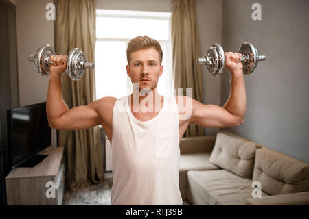 Young well-built man go in for sports in apartment. He doing shoulder press using dumbbells. Guy look straight forward. Serious muscular and strong Stock Photo