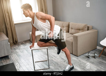 Young well-built man go in for sports in apartment. He does dumbbell row exercise. Concentrated strong muscled young man Stock Photo