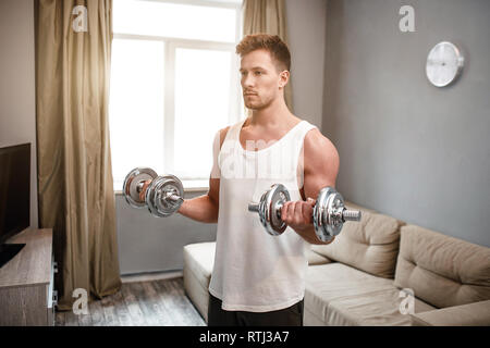 Young well-built man go in for sports in apartment. Guy doing biceps dumbbell exercise with both hands. Serious concentrated young man with muscular b Stock Photo