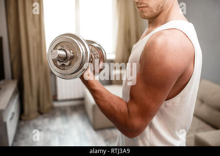 Young well-built man go in for sports in apartment. Cut view of guy doing biceps dumbbells exercises alone. He look at muscles. Daylight Stock Photo
