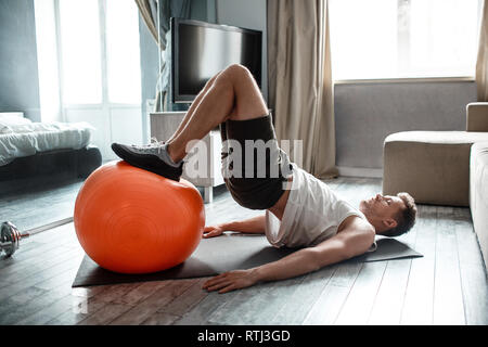 Young well-built man go in for sports in apartment. Guy hold legs on fitness ball and doing lifting up with body. Strong hardworking man Stock Photo