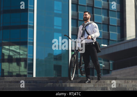 Enjoying beautiful cityview. Businessman standing on stairs with bicycle, having break in riding, copy space Stock Photo