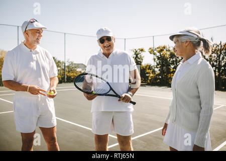 Cheerful men in tennis wear talking to a senior woman standing on a tennis court. Smiling old man standing with his playmates holding a tennis racket. Stock Photo