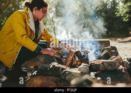 Man putting fire wood in a bonfire in a forest. Man camping in a countryside location setting up a campfire. Stock Photo