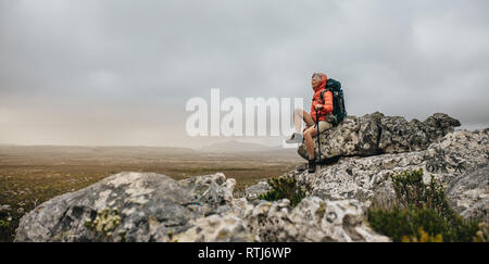 Senior woman sitting on a rock enjoying the scenic view from the top. Woman trekker relaxing after finishing her trek on a hill. Stock Photo