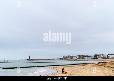 Woman walking her dog in foreground on Margate sands in mid-winter under overcast grey sky. Town, main sands and harbour in background. Negative space Stock Photo