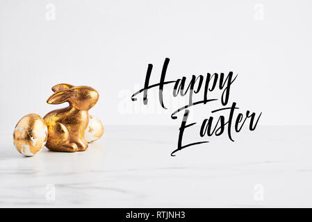 Easter greeting card, golden rabbit and eggs decorated with golden foil on white marble table with quote Happy Easter. Stock Photo