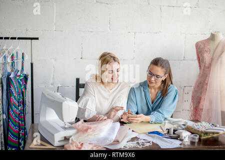 Confident seamstress are discussing clothes models with interested beautiful female client, sitting at working place and looking at smartphone Stock Photo