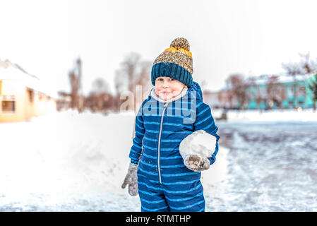 A little boy 3-6 years old blue jumpsuit is standing in hand of big snowball. Winter in city on background of snowdrifts of snow and trees. Happy