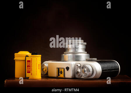 Horizontal shot of a vintage 35mm camera and film with yellow metal canister on a brown background with copy space.  The camera is lying on its back w Stock Photo