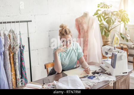 Creative female seamstress in eyeglasses and tape working on design of new garment drawing sketches sitting at desktop in vintage stylish atelier Stock Photo