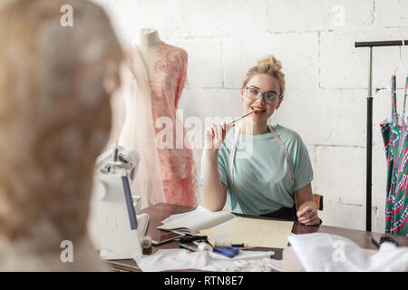 Dreamy thoughtful seamstress with blonde hairmaking notes in sewing studio in soft pink and mint tones, making a work plan for the next week Stock Photo