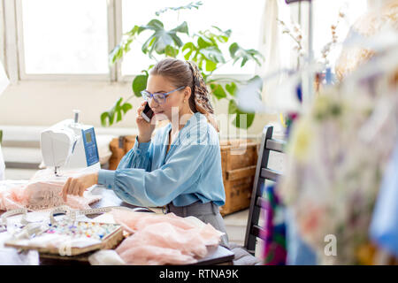 Young female fashion designer in blue elegant blouse is taking the order, discussing bespoke party dress with customer by phone Stock Photo