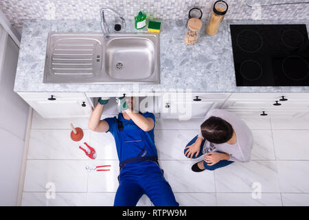 High Angle View Of Woman Looking At Handyman Lying On Floor Repairing Sink In Kitchen Stock Photo