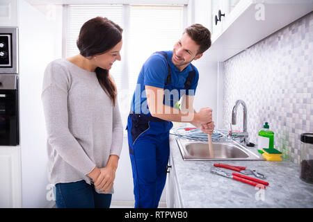 Happy Woman Looking At Male Plumber Using Plunger In The Kitchen Sink Stock Photo