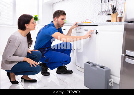 Serviceman Crouching On Kitchen Floor Pressing Button Of Dishwasher While Woman Looking At Him Stock Photo