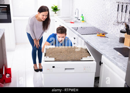 Smiling Woman Behind Technician Repairing Dishwasher In Kitchen Stock Photo