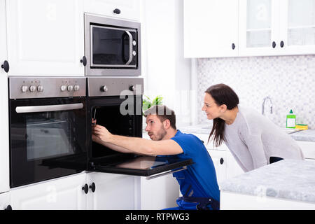 Smiling Young Woman Looking At Technician Fixing Oven In Kitchen Stock Photo