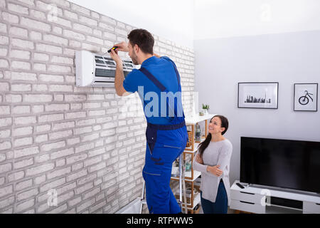 Smiling Woman Looking At Male Technician Repairing Air Conditioner With Screwdriver In Home Stock Photo
