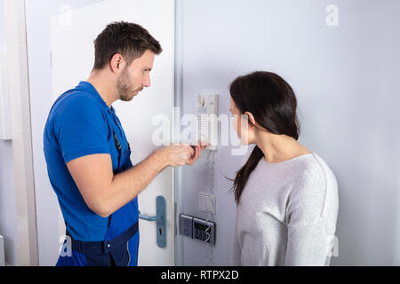 Woman Standing In Front Of Door While Repairman Fixing An Intercom Phone With Screwdriver Stock Photo