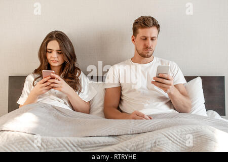 Young couple in the bed after waking up. An attractive woman and man are checking social networks news in mobile phones, texting or sending messages Stock Photo