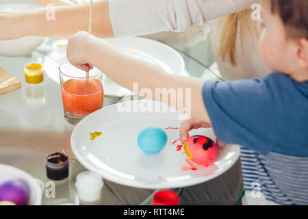 Preschooler boy helping his mother to paint out the boiled white eggs for Easter Holiday Celebration, focus on hand, dipping the paintbrush in a glass Stock Photo