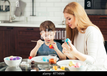 happy blonde mother and four-year old son making homemade Easter eggs, enjoying leisure time spent together at home. Family celebrating Easter. Parent Stock Photo