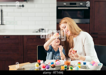 Young beautiful blonde mother and her cute four year old son having fun while painting eggs for Easter at home kitchen, smiling, developing creative a Stock Photo