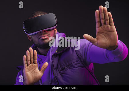 Handsome afro american man wearing VR glasses while working over virtual project, posing isolated against dark wall with copy space. Smart innovative  Stock Photo
