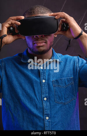 Handsome african designer in denim wearing VR glasses while working over virtual project, posing isolated against dark wall with copy space. Smart inn Stock Photo