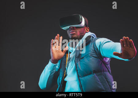 Gaming concept. African casual man in warm seasonal outdoor clothes wearing mobile VR headset enjoying new reality in dark room. Fashion and Innovatio Stock Photo