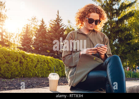 Attractive young girl student with curly red hair having coffee and using smartphone in summer park. Stylish woman chilling outdoors Stock Photo