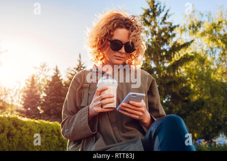 Hipster young woman with curly red hair drinking coffee and using smartphone in summer park. Stylish girl chilling outdoors Stock Photo